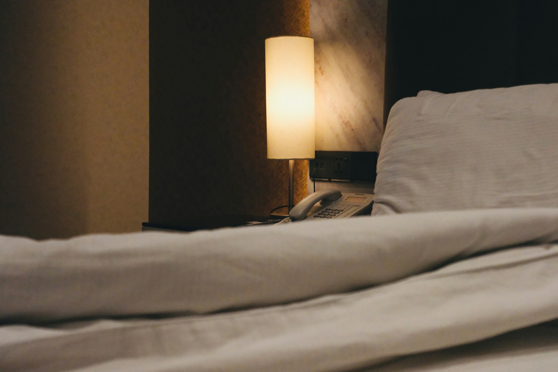 A dimly lit bedside lamp illuminates a rumpled white duvet.